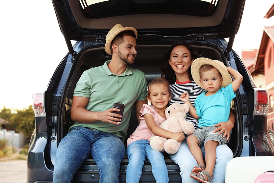 About Our Agency - Parents and Their Small Children Sit in the Tailgate of Their Car in Front of Their Home, Smiling and Holding a Teddy Bear